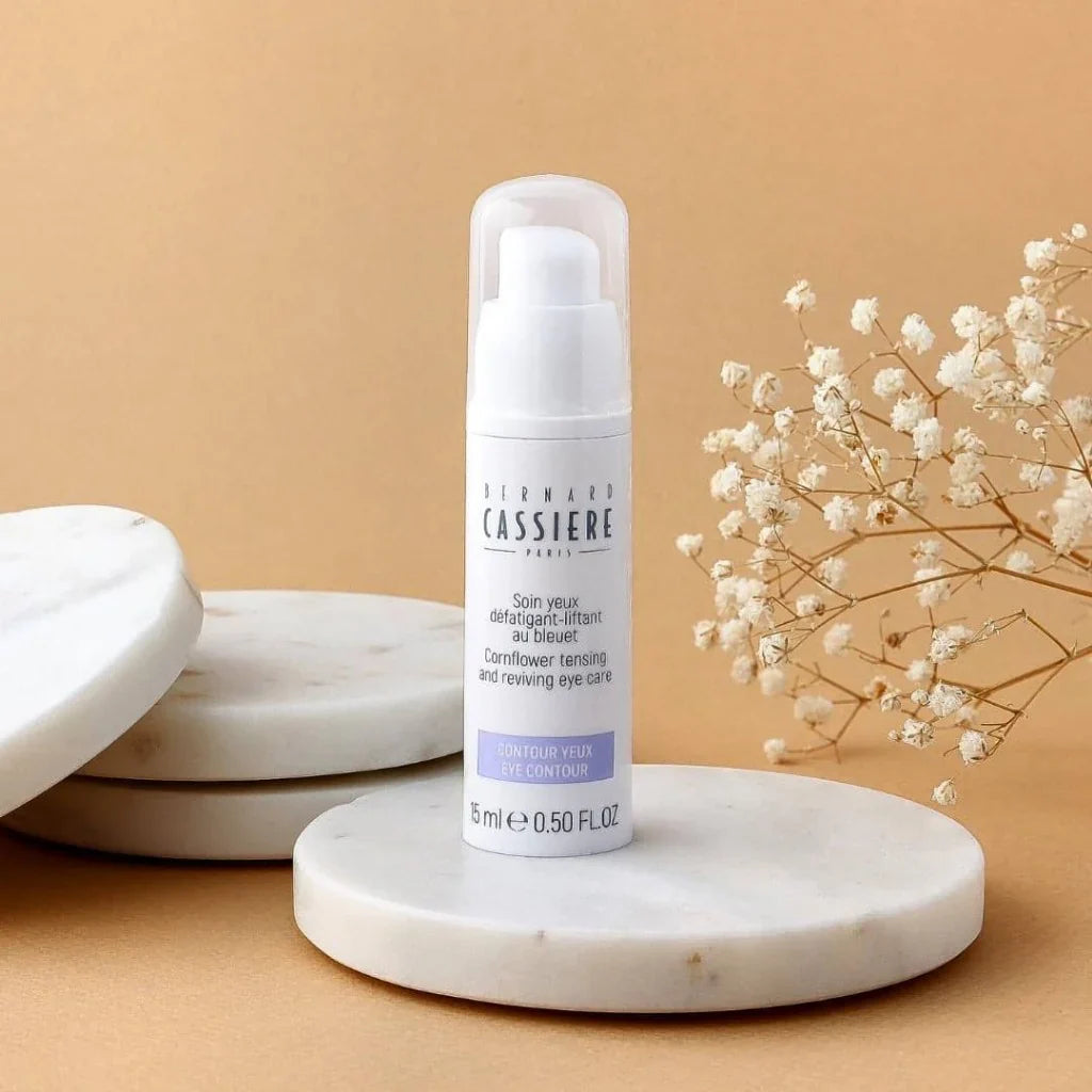 A bottle of Bernard Cassiere Paris Eye Contour eye cream, displayed on a white marble coaster against a warm beige background with dried baby's breath flowers.