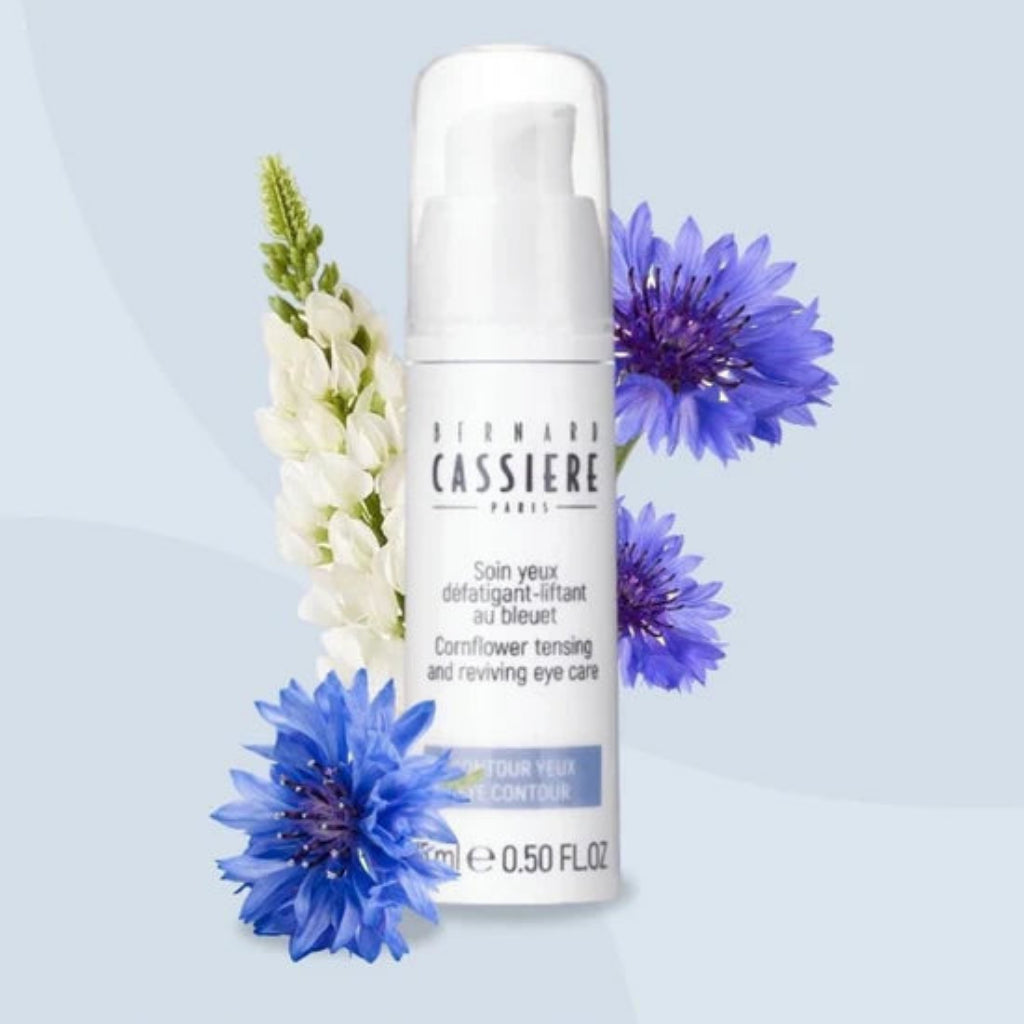 A white pump bottle of Bernard Cassiere Paris eye care cream, styled with blue cornflowers and white flowers against a pale blue background.