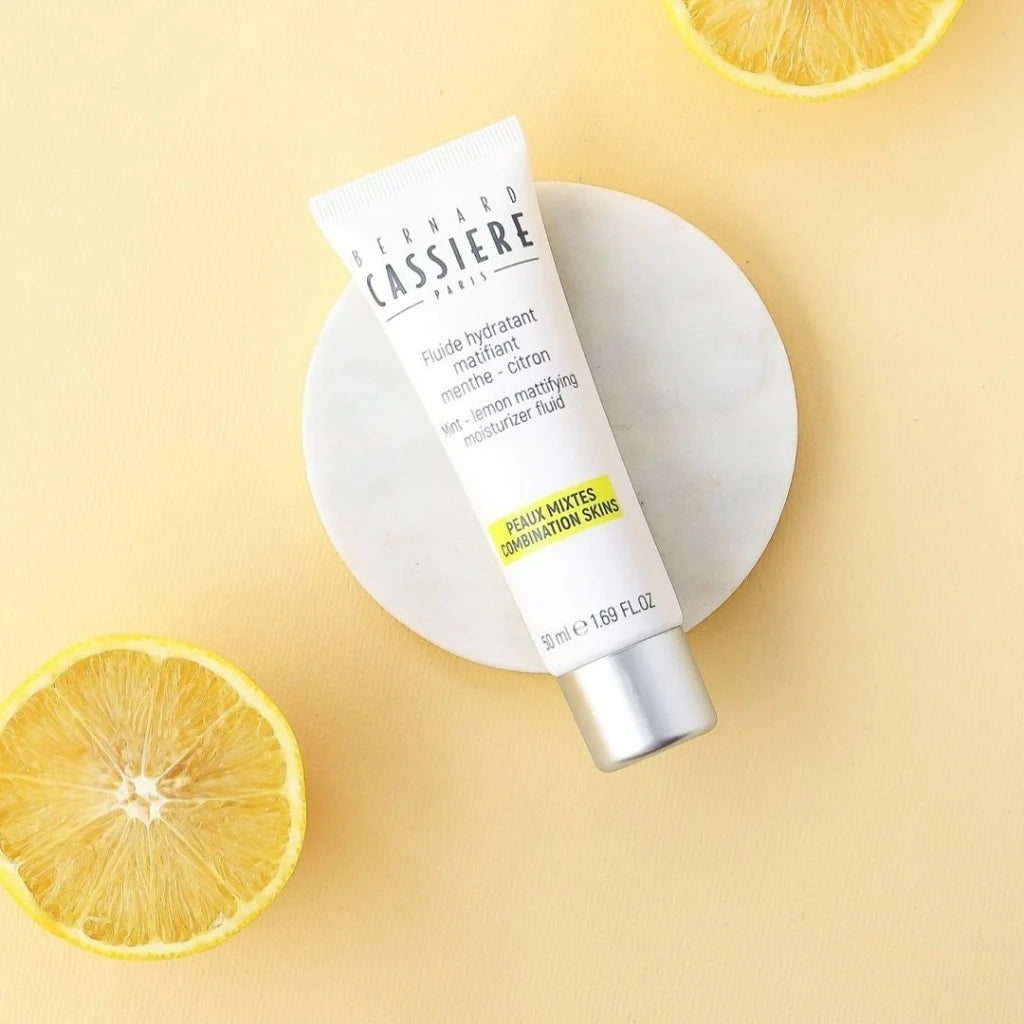 A squeeze tube of Bernard Cassiere lemon mattifying moisture fluid set on a small white round plate, styled with lemon slices on a pale yellow background.
