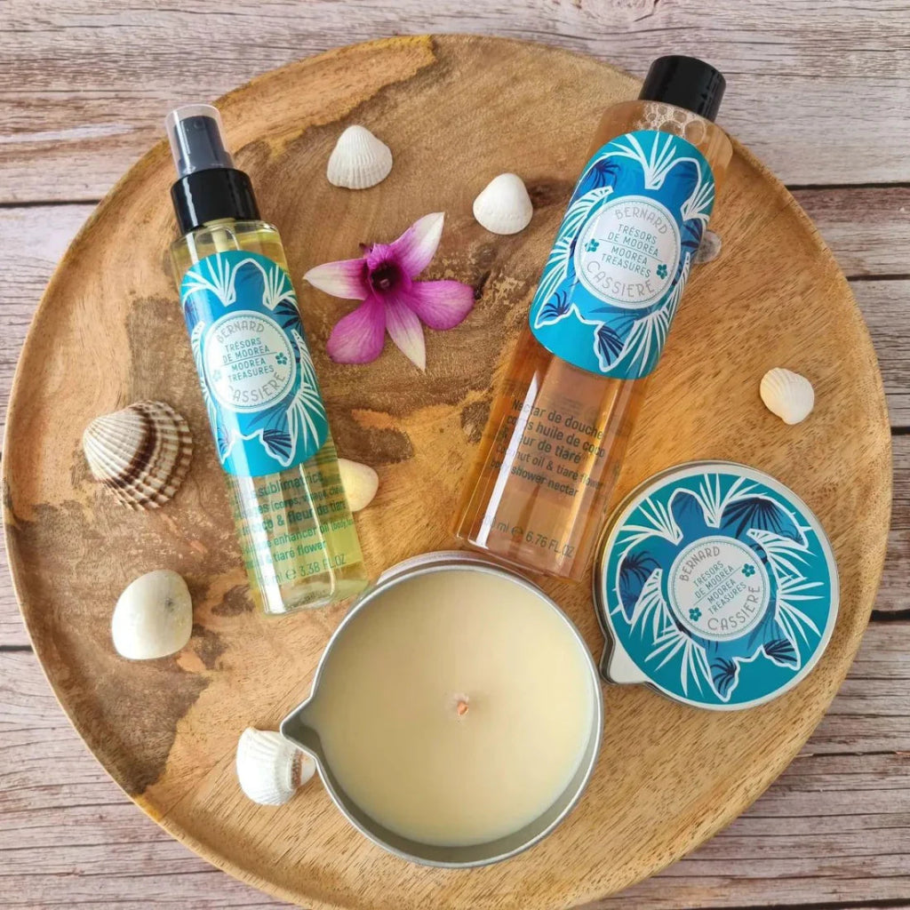 A tropical-themed bath and body set arranged on a round wooden tray, featuring a body oil, a shower nectar, a candle, and a matching tin lid, styled with seashells and a small purple flower on a rustic wood background.