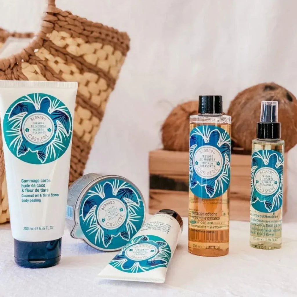 A set of Coconut and Tiare Body Care products, including an exfoliant, massage oil candle, body lotion and hair and body oil, styled with a woven bag and coconuts on a white surface.