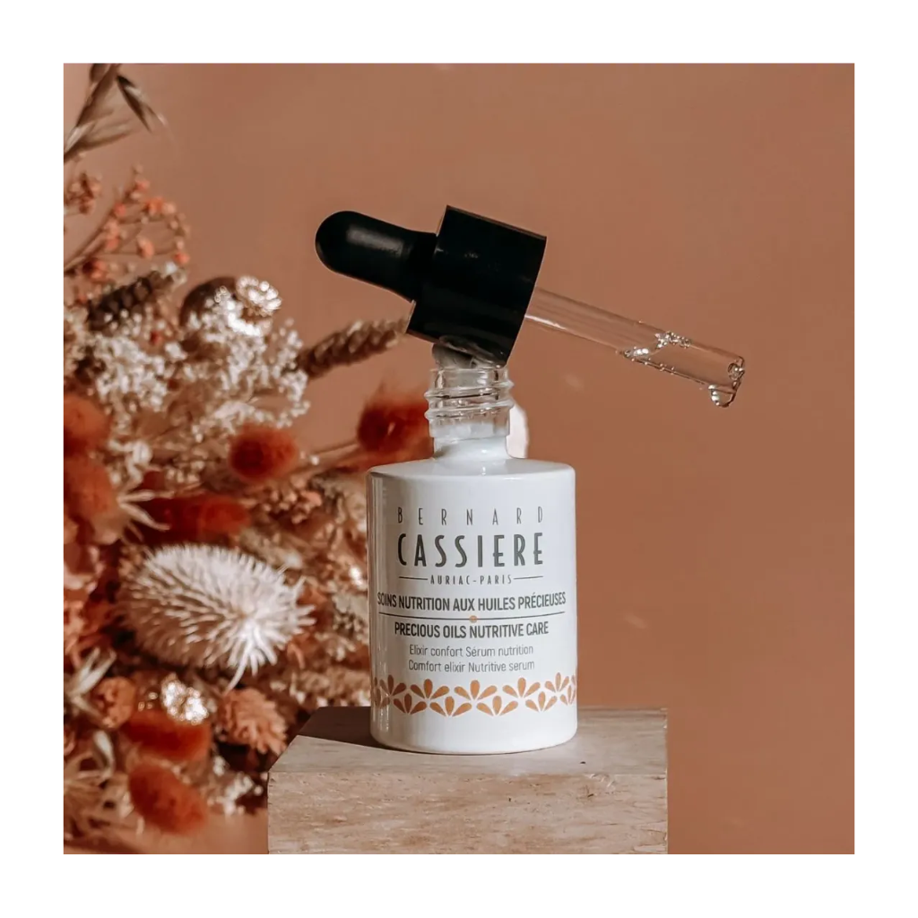 A bottle of Bernard Cassiere comfort elixir nutritive serum with a black dropper cap, displayed on a wooden block next to dried flowers against a warm terracotta background.