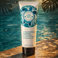 A tube of Bernard Cassiere beautifying body milk standing upright on a poolside surface with a swimming pool and palm leaves in the background