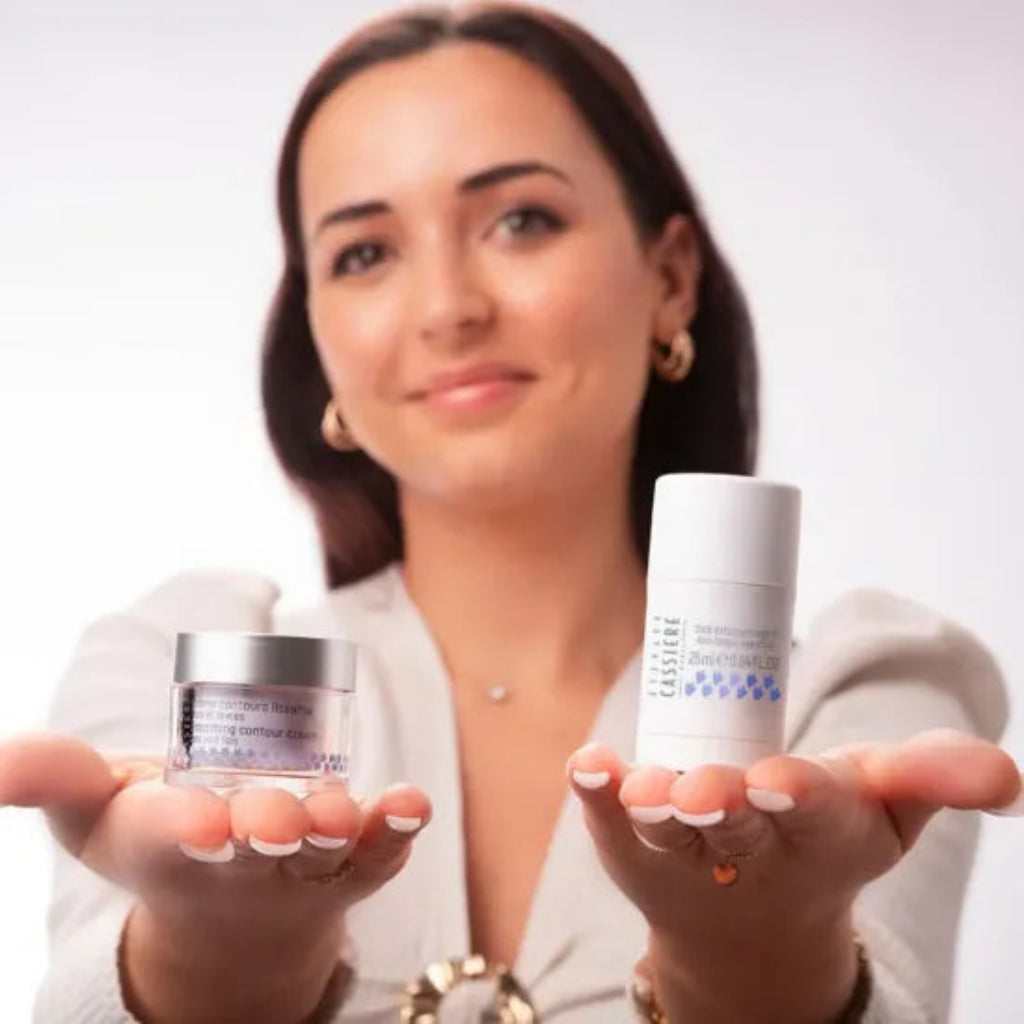 A woman holds up two Bernard Cassiere eye products in her hands against a clean white background. On the left, she holds a small jar of the smoothing contour cream, on the right she holds the anti-fatigue eye stick.
