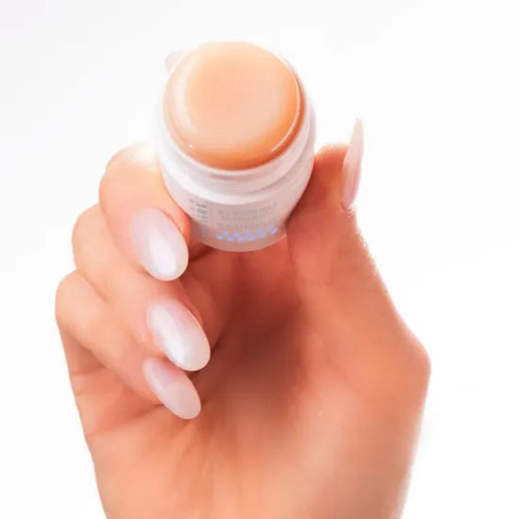 A hand with long, clean, off-white manicured nails holds the Bernard Cassiere anti-fatigue stick, a peach-colored cream against a plain white background.