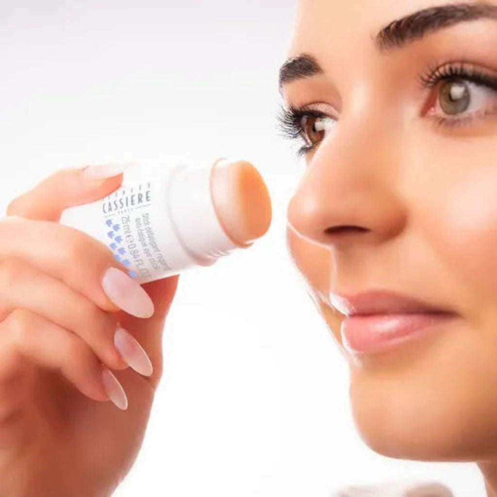 A close-up of a woman holding a stick of Bernard Cassiere anti-fatigue stick near her eye against a bright white background.