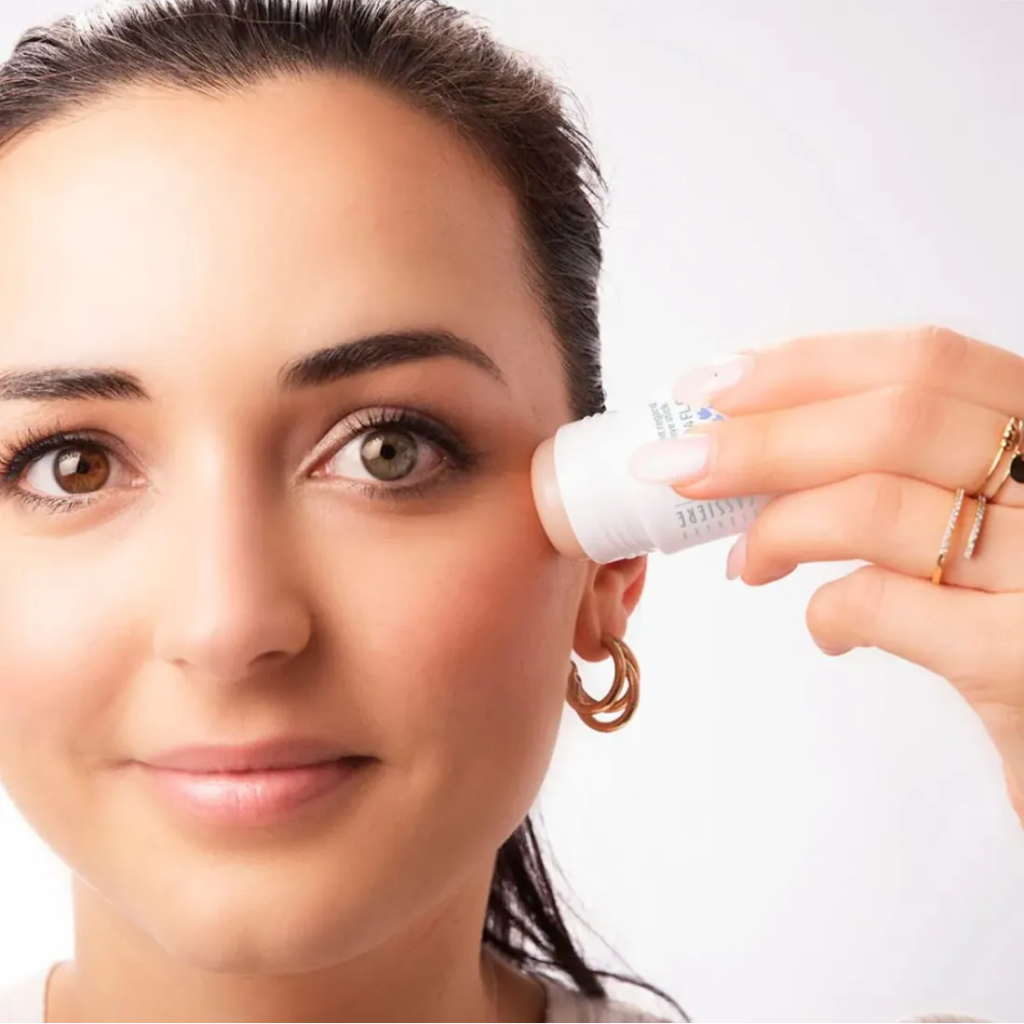 A close-up of a woman holding a stick of Bernard Cassiere anti-fatigue stick near her eye against a bright white background.