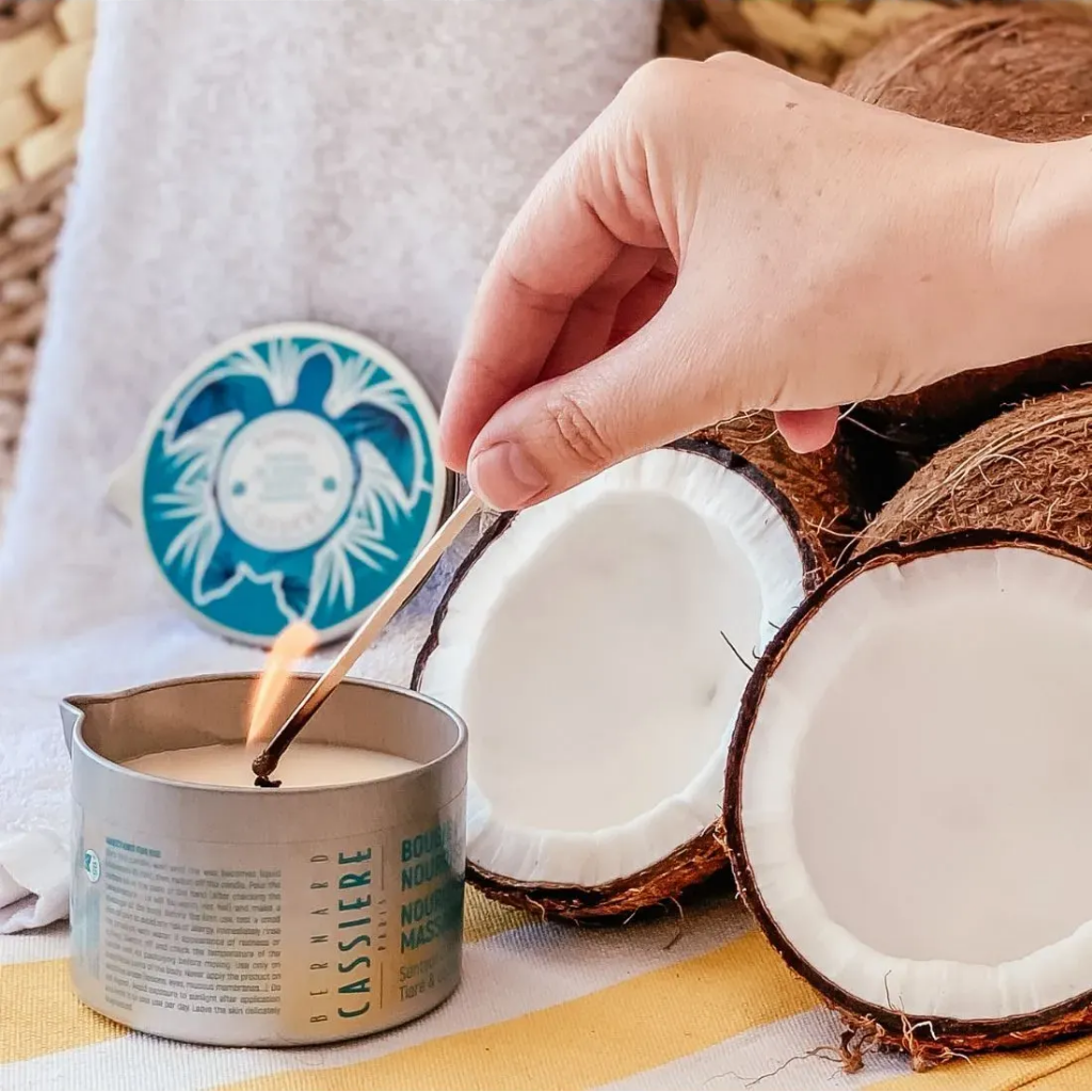 A hand lighting a small metal candle tin with a wooden match. The candle tin is styled with halved coconuts, a towel, and a round blue-and-white decorative lid in the background.
