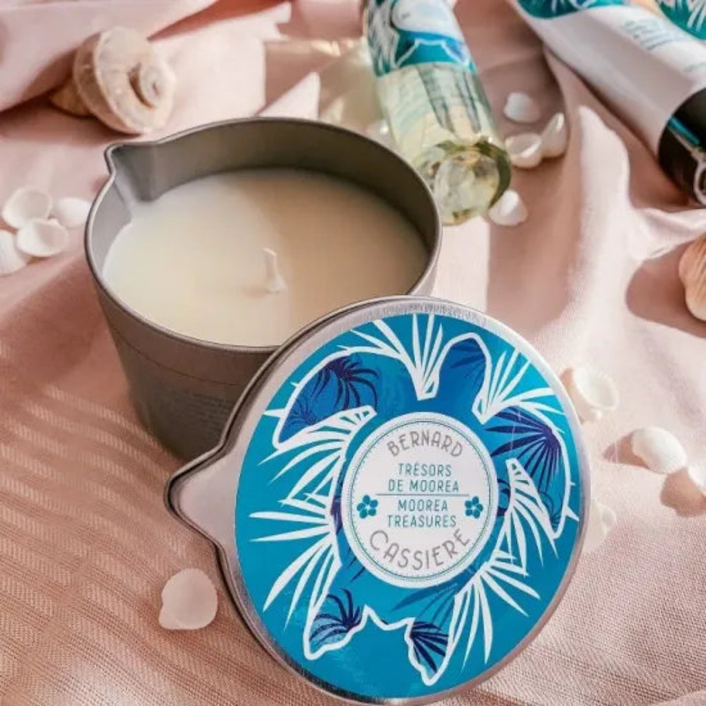 An open metal candle tin with creamy off-white candle wax beside its round lid, which has a blue tropical leaf design. The scene is styled on pink fabric with seashells and a matching bottle in the background.