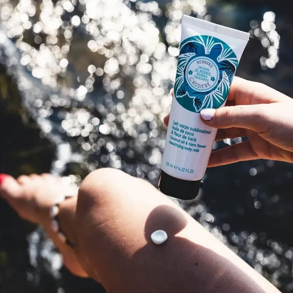 A hand holds a tube of the bernard cassiere coconut oil and tiare flower beautifying body milk in front of a their leg with a dollop of the white cream on it, set against a blurred outdoor background of sparkling water.