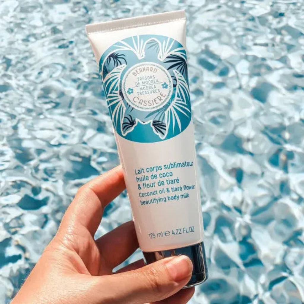 A hand holds a tube of Bernard Cassiere beautifying body milk against a background of rippling blue water.