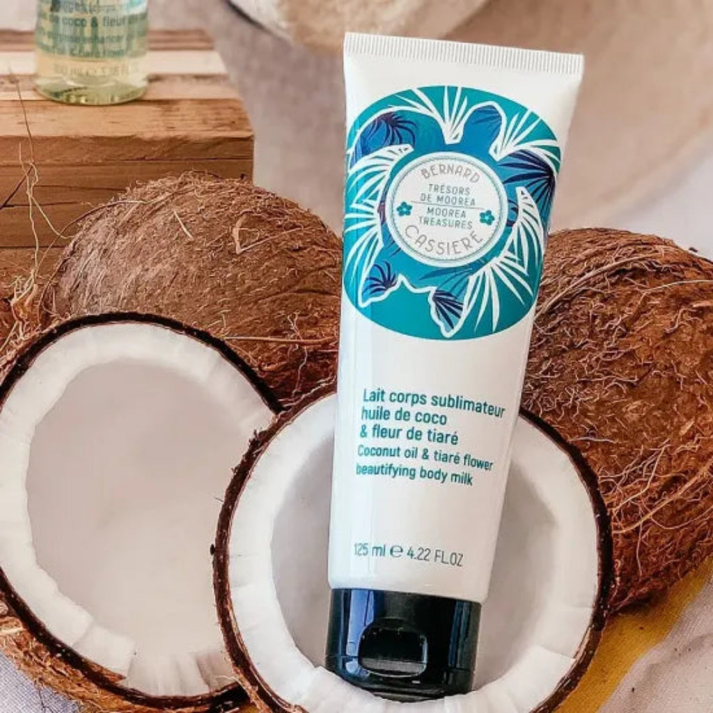 A squeeze tube of Bernard Cassiere Coconut Oil & Tiare Flower Body Milk stands upright in the center, surrounded by halved coconuts on a rustic wooden surface.