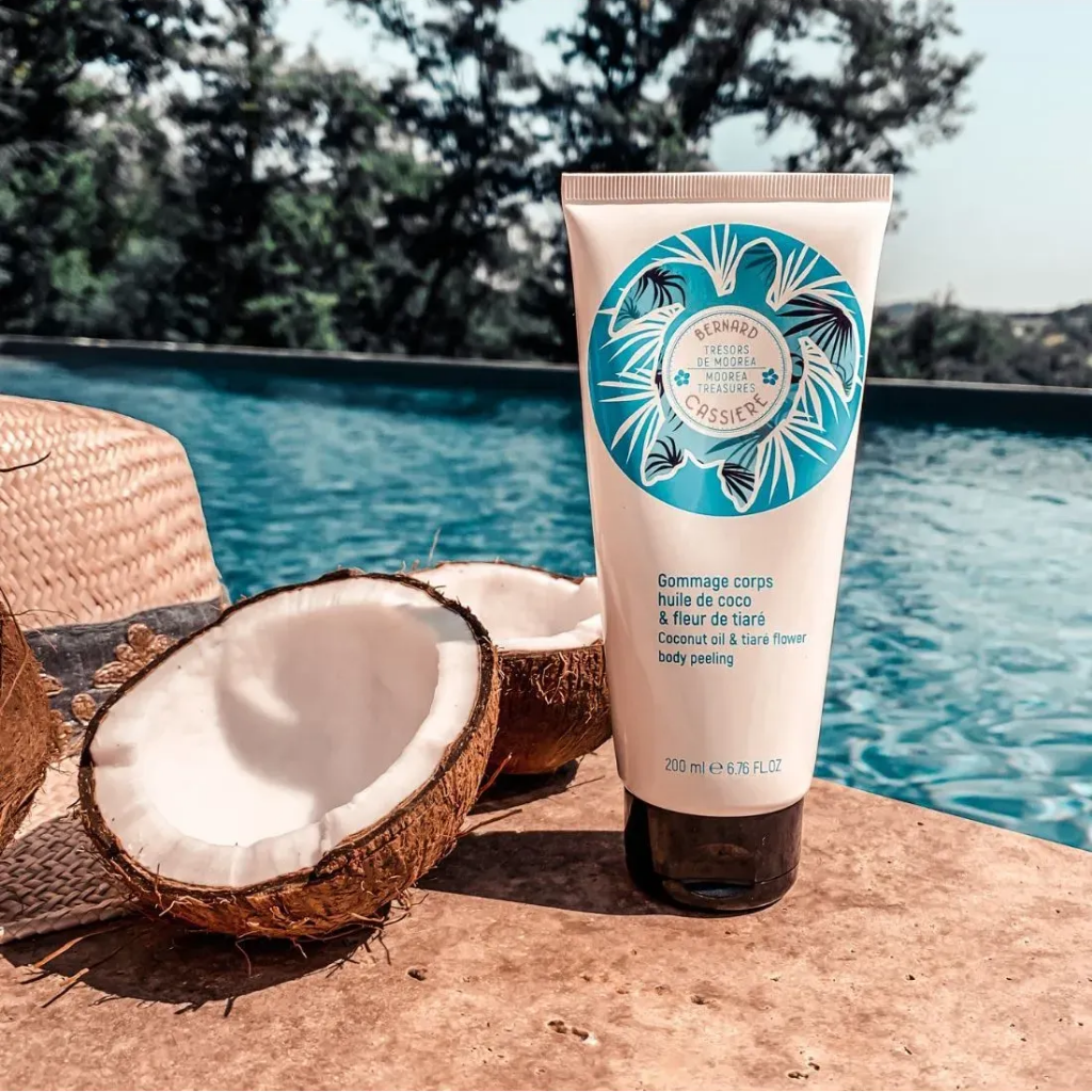 A tube Bernard Cassiere coconut oil and tiare flower body peeling stands on a stone surface beside halved coconuts, with a swimming pool and trees in the background.
