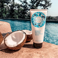 A tube Bernard Cassiere coconut oil and tiare flower body peeling stands on a stone surface beside halved coconuts, with a swimming pool and trees in the background.