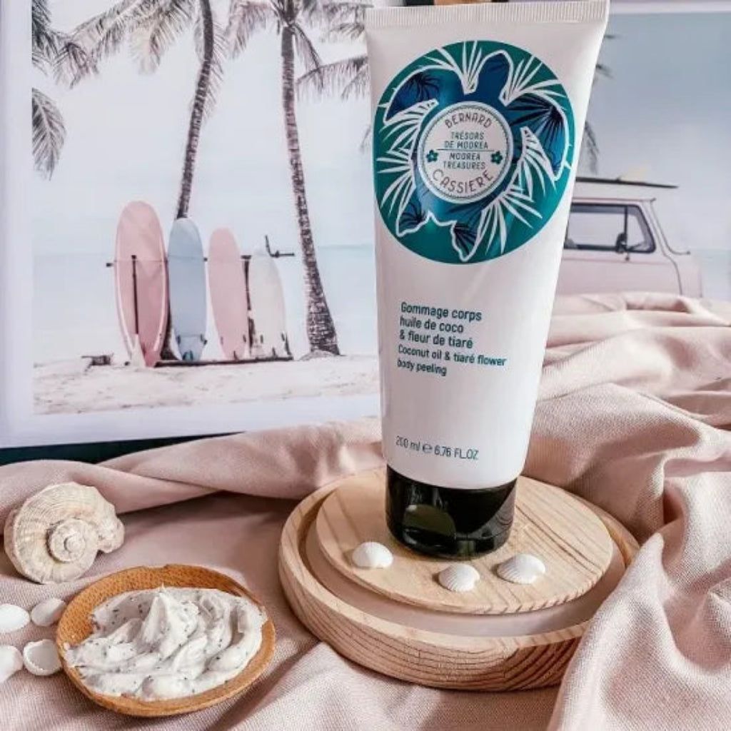 A tube Bernard Cassiere coconut oil and tiare flower body peeling stands on a wooden tray, surrounded by seashells, a small dish of textured cream, and a beach-themed print in the background.