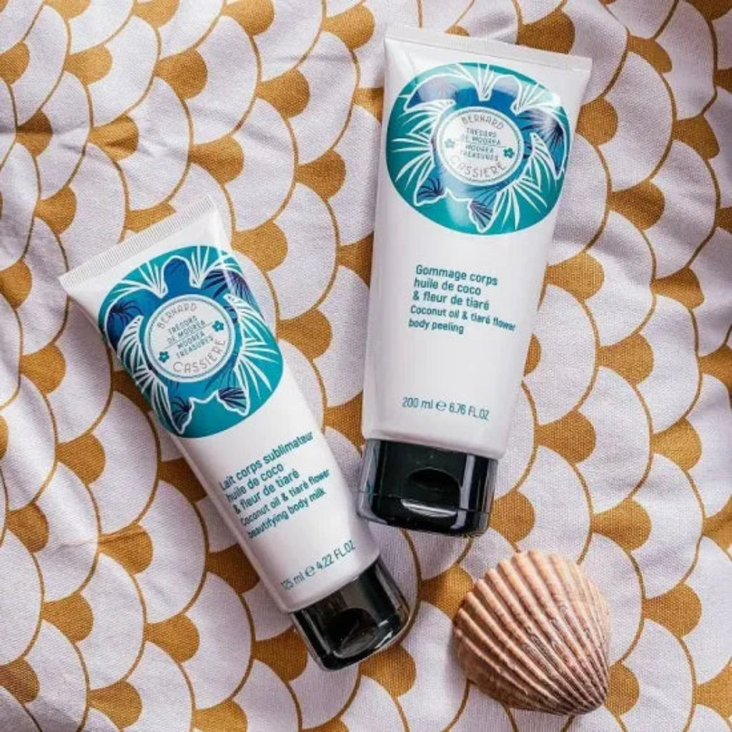 Two tubes of Bernard Cassiere coconut oil & tiare flower body care, one body milk and one body peeling, arranged on a patterned white and gold towel next to a seashell.