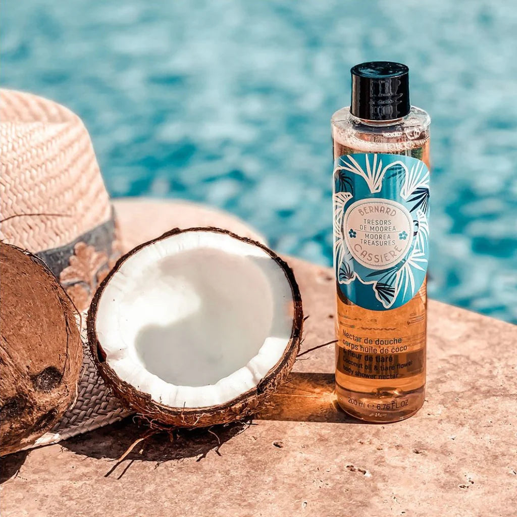 A bottle of Bernard Cassiere Coconut & Tiare body shower nectar stands on a stone surface beside a halved coconut, a woven hat, and a blurred turquoise pool background.