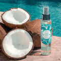 Multi-purpose enhancer oil pump bottle next to halved coconuts in front of turqouise water