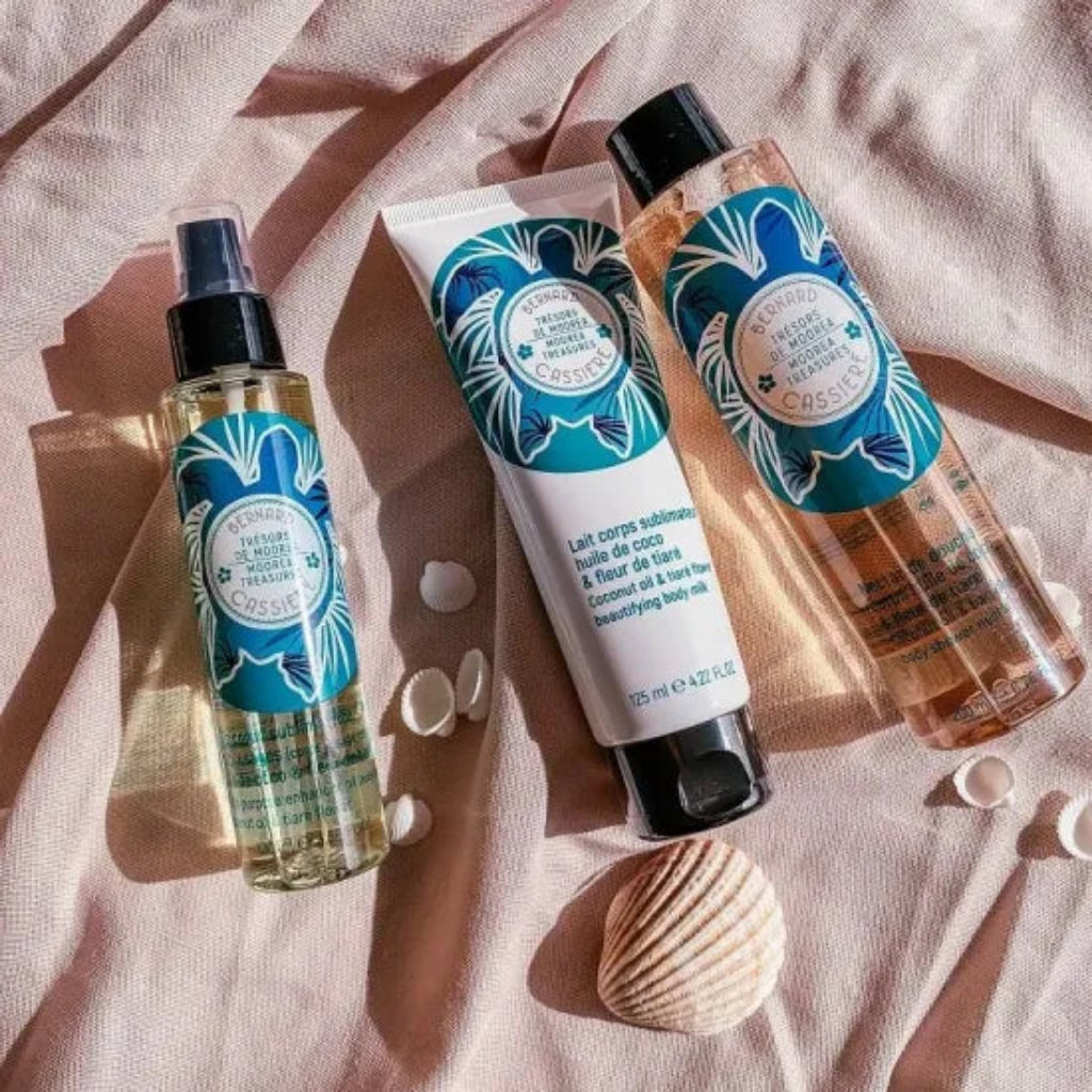 Three Bernard Cassiere coconut oil and tiare flower body care products arranged on draped blush pink fabric with small seashell props: multi-purpose enhancer oil (a clear pump bottle), beautifying body milk in a squeeze tube, and shower nectar, (body wash) a peach-tinted liquid 