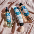 Three Bernard Cassiere coconut oil and tiare flower body care products arranged on draped blush pink fabric with small seashell props: multi-purpose enhancer oil (a clear pump bottle), beautifying body milk in a squeeze tube, and shower nectar, (body wash) a peach-tinted liquid 