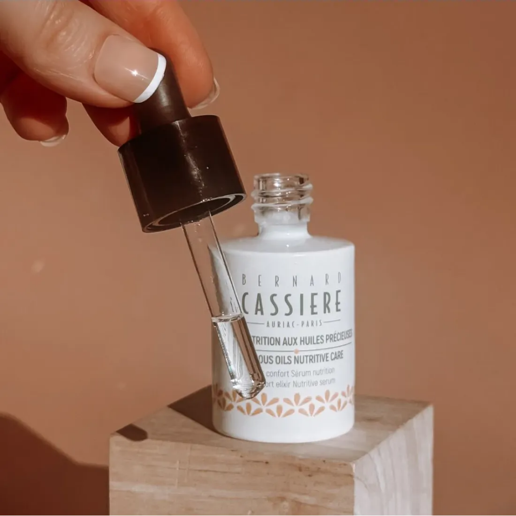 A hand with a French manicure holds a black dropper above an open white bottle of Bernard Cassiere comfort elixir Nutritive Care serum, set on a wooden block against a warm beige background.