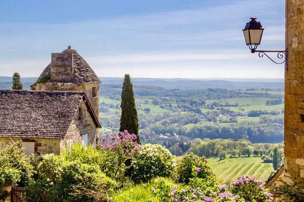 A scenic landscape photograph of a stone village with a rustic house and old-fashioned wall lamp overlooking rolling green hills under a blue sky.