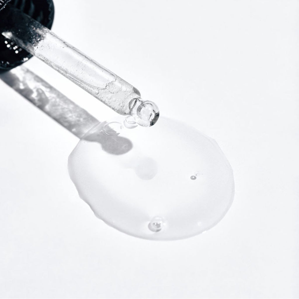 A close-up of a clear glass dropper above a glossy pool of transparent serum on a white background.