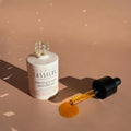 A bottle of Bernard Cassiere Healthy Glow-Boosting Elixir with its dropper lying beside a small pool of golden-yellow serum on a warm beige surface.