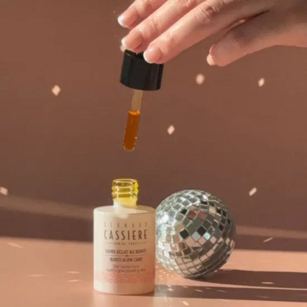 A hand holds a black-capped dropper above an open bottle of Bernard Cassiere healthy glow-boosting elixir, with amber liquid dripping into the bottle. A mirrored disco ball sits beside the bottle on a warm brown surface against a matching brown background.