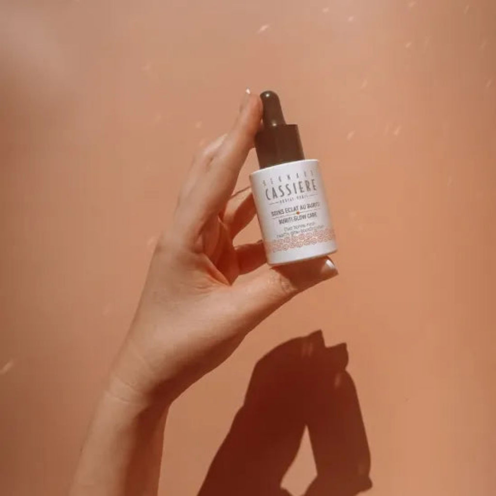 A hand holding a white dropper bottle of Bernard Cassiere Healthy Glow-Boosting Elixir Serum against a warm peach-colored background with a shadow.