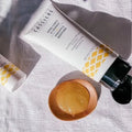 Bernard Cassière Pineapple silhouette body peeling tube in white with yellow diamond pattern, shown on a textured white fabric alongside a small wooden dish with the light yellow gel inside.