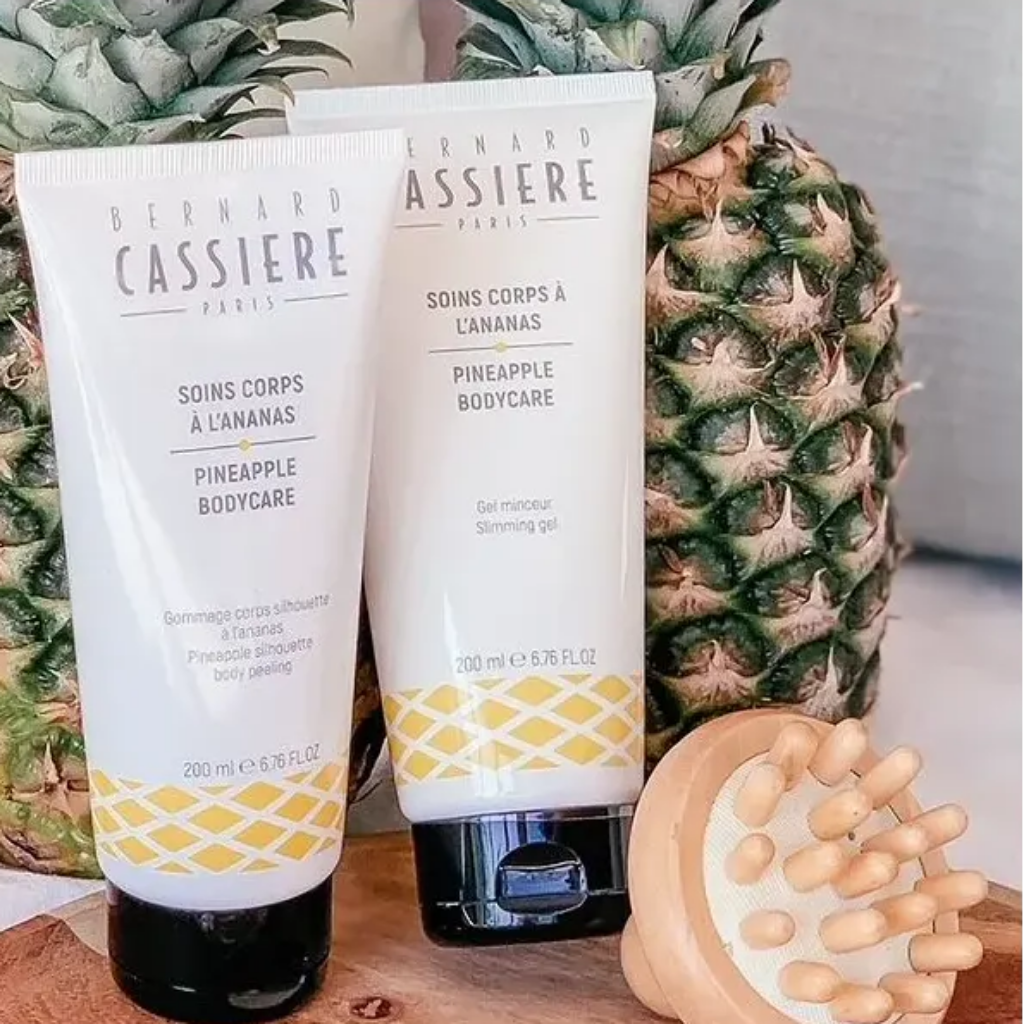 Two tubes of Bernard Cassière body products with pineapple graphics, displayed in front of whole pineapples and a wooden body tool.