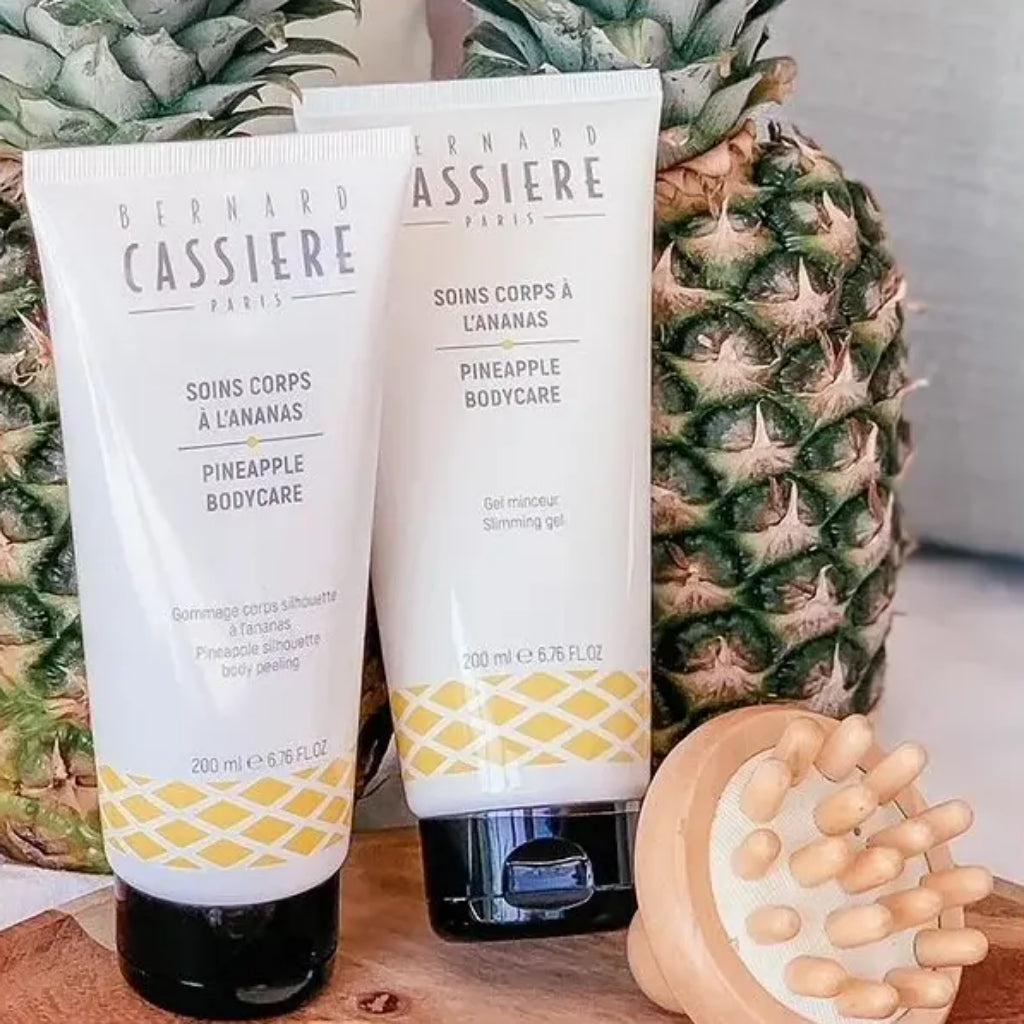 Two tubes of Bernard Cassière body products with pineapple graphics, displayed in front of whole pineapples and a wooden body tool.
