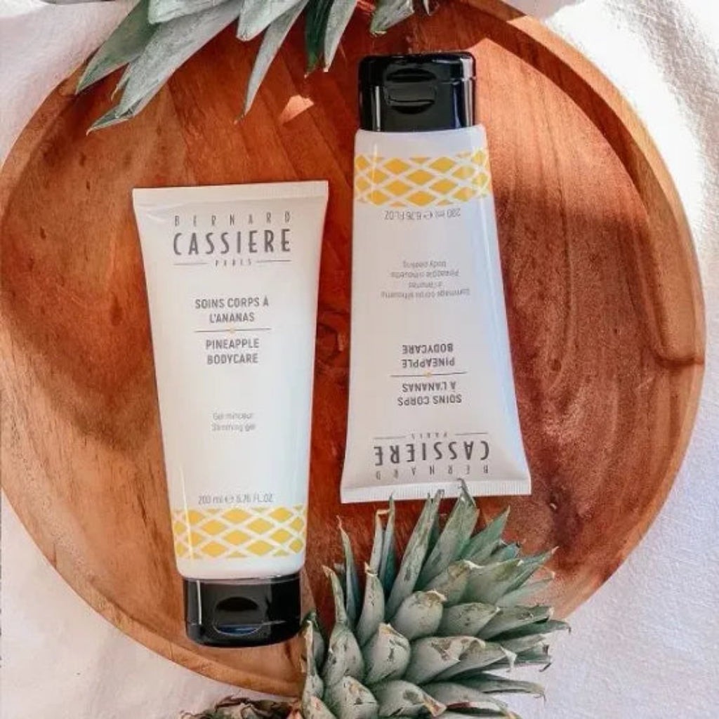 Two tubes from Bernard Cassiere Paris Pineapple Bodycare on a round wooden tray, styled with pineapple leaves. The products are a slimming gel and a body peeling, both white with yellow geometric patterns and black caps.