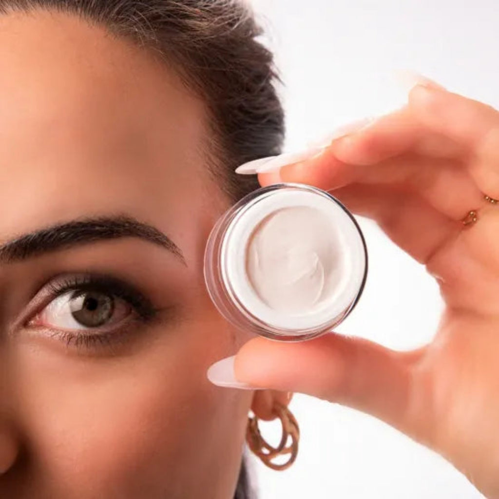 A close-up lifestyle shot of a woman holding a jar of Bernard Cassiere eye and lip smoothing contour cream jar near her eye, showcasing the product texture against a clean white background.